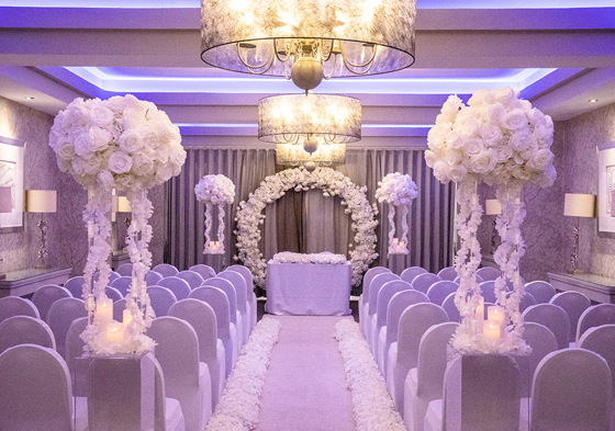 White ceremony room with purple-hued lighting and white rose arch and bouquets